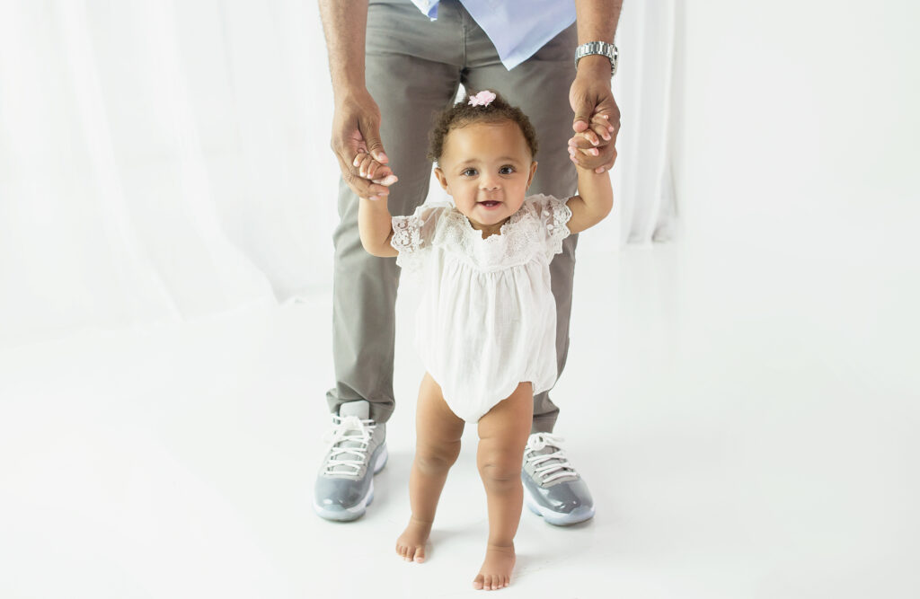 Little girl wearing white romper, holding dads hands during first birthday cake smash photoshoot in Franklin Tennessee photography studio 