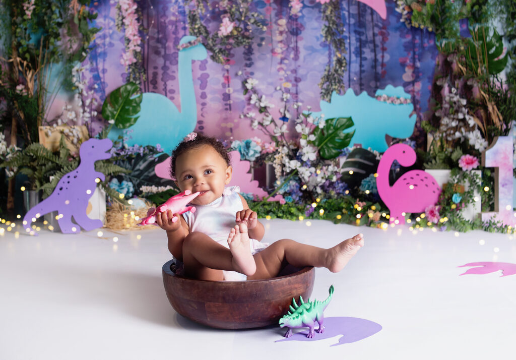 Baby girl sitting in bowl holding dinosaur toy laughing during first birthday cake smash session in Mount Juliet Tennessee photography studio 